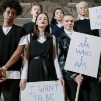A group of individuals in dark clothing and makeup holding signs that say, "I am who I am".