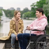 Talk therapy being conducted by an individual in a wheelchair and another individual sitting on a bench next to water.